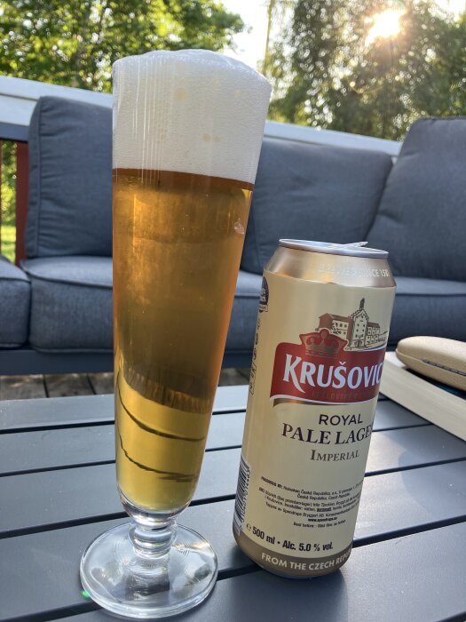 Högt glas med skummande öl bredvid en burk Krusovice Royal Pale Lager på ett trädgårdsbord med soffa och solsken i bakgrunden.