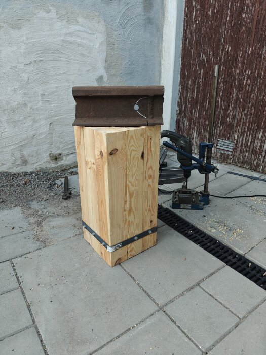 Träpelare med metallbeslag står på en trottoar intill ojämn vägg, metallverktyg i bakgrunden.