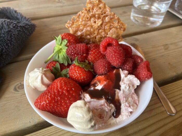 Skål med jordgubbar, hallon, maränger, knäckflarn och grädde med chokladsås på träbord.