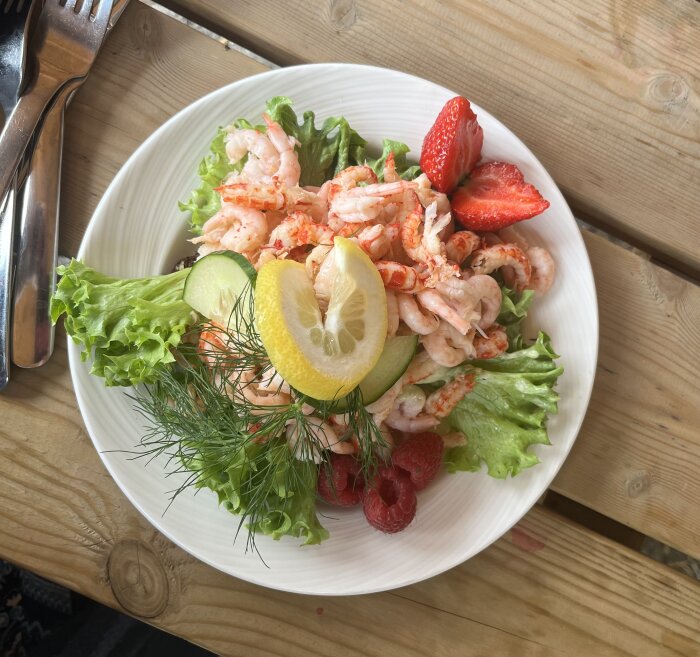 Kräft- och räkmacka med sallad, citron, dill, skivad gurka och jordgubbar serverad på tallrik på träbord.