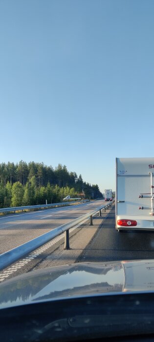 Kö av bilar på motorvägen med husvagn framför; skog i bakgrunden och klarblå himmel. Emojin antyder frustration.