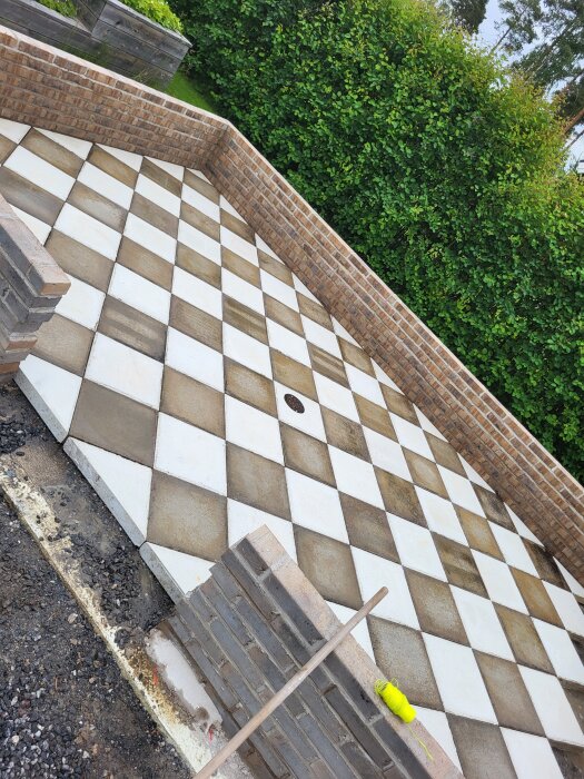 Sockel av tegel med schackrutigt golv förberett för orangeri, omgiven av grön häck. Stödmur på ena sidan med verktyg framme.