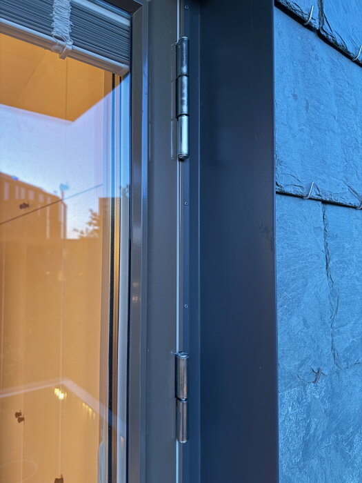 Närbild på dörrgångjärn med justeringsmöjligheter på insexskruvar mot en grå vägg och fönster, del av tätning synlig.