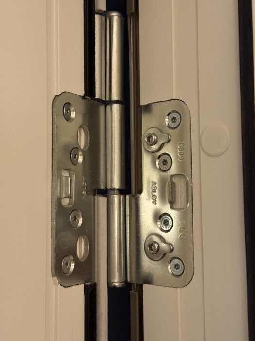 Närbild på ett gångjärn med synliga skruvar, monterat på en dörrkarm. Gångjärnet kan justeras med insexnyckel.