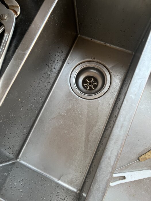 Rostfri diskho med utlopp och lite vattenstänk, en tång syns i bakgrunden. Diskhon är från 2008 och har problem med läckage i utloppet.