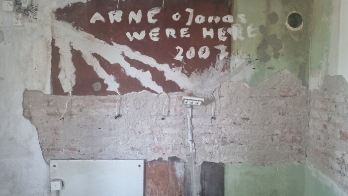 Renoveringsobjekt med synlig tegelvägg där putsen har lossnat, texten "Arne och Jonas were here 2007" syns på väggen, elektriska kablar löper längs ytan.