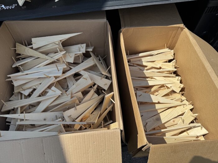 Två kartonger fyllda med träkilar eller tältpinnar för kokosmatta eller markduk, upplagda för försäljning.