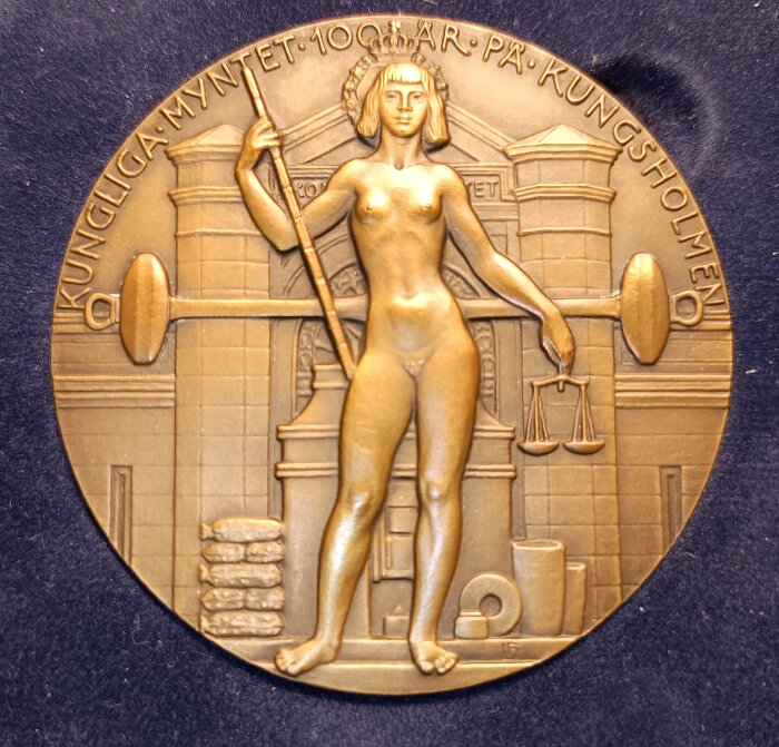 Medalj i brons med en kvinna som håller våg och scepter, omgiven av texten "Kungliga Myntet 100 år på Kungsholmen".