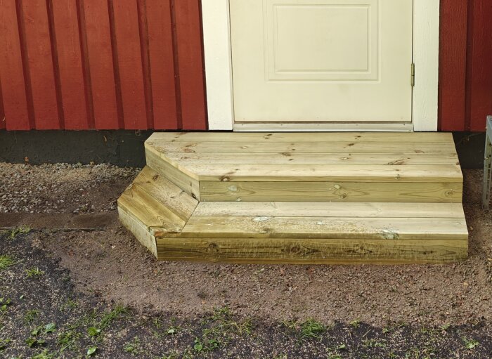 Liten trappa av tryckt virke vid en röd vägg och vit dörr, byggd på sand. Övervägande trädgårdsplattor istället för ökad hållbarhet.