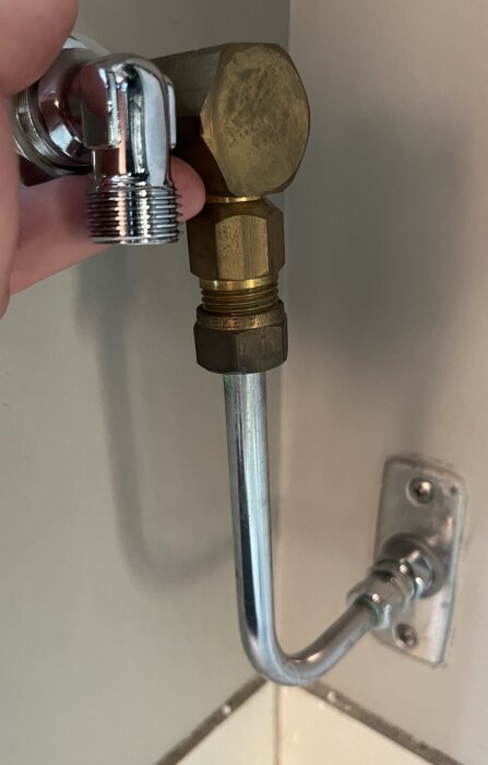 Hand håller i kromad koppling bredvid vinklad mässingskoppling ansluten till vägg och rör i badrum eller kök.