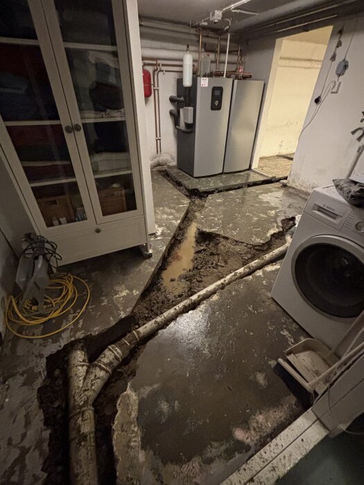 Badrumsrenovering i källare med uppbruten betong och synliga rör, tvättmaskin och pannrum i bakgrunden, redo för avloppsinstallation.