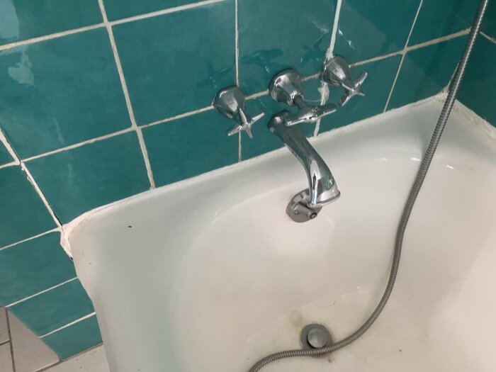 Badkar med blå kakelplattor, tre kranar och en duschslang.