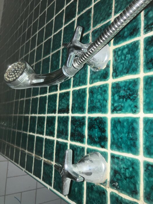 Duschblandare med handdusch och två vred monterade på grönkaklad vägg.
