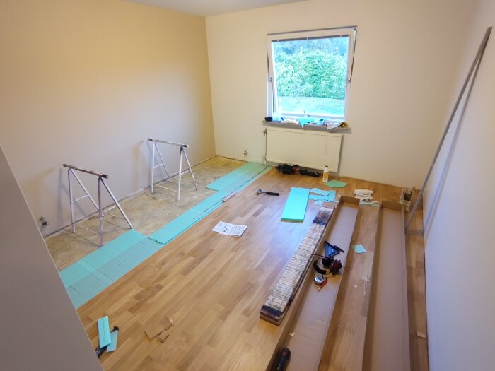 Rum under renovering med nymålade väggar och halvlagt klickgolv. Verktyg och material ligger utspridda på golvet, samt kartonger och golvfoam.