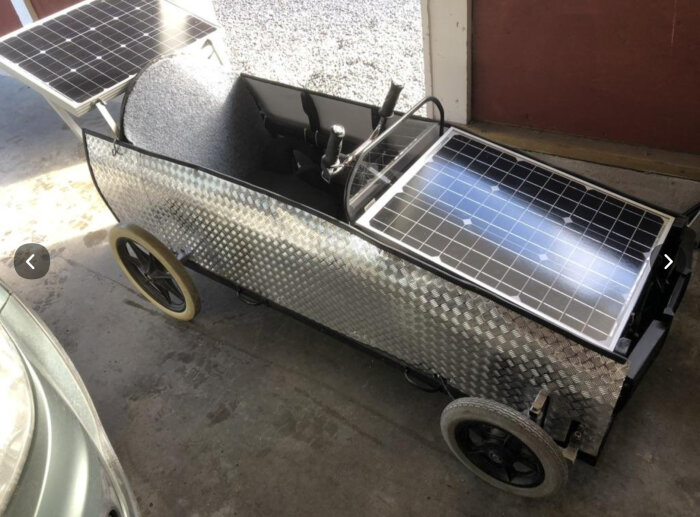 Liten bil täckt med metallplåt och solpaneler, modifierad med elmotor, visas i ett garage.