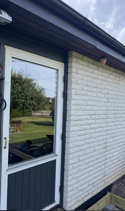 Murad fasad med vit tegel, svart hängränna, en dörr till vänster och trädgård i bakgrunden under klar himmel.