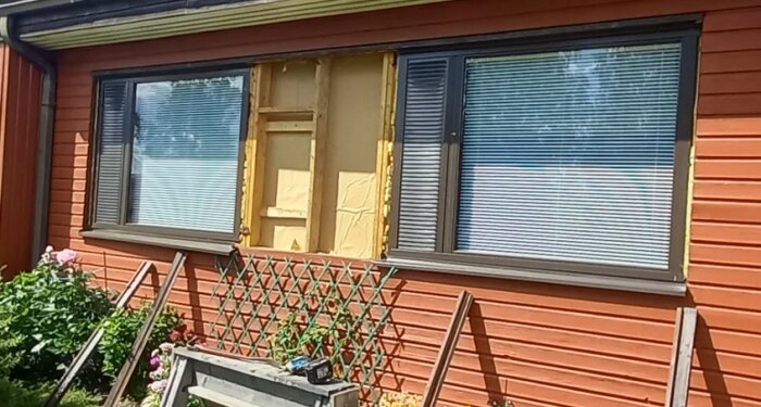 Renoverad fasad med ny isolering och panel, samt färdigt fönster med jalusier. Dubbla fönster och trädgårdsdetaljer synliga i förgrunden.