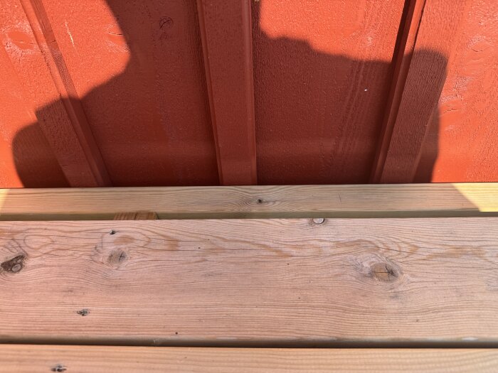 Puuterassi punaisen talon seinän edessä, puun syyt selvästi näkyvissä, varjo peittää osan seinästä.