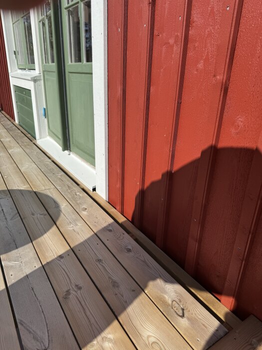 Punainen seinä ja vihreä ovi näkyvät kuistilta. Terassi on osittain valmis, laudat asennettu vierekkäin seinän viereen, varjot näkyvät laudalla.