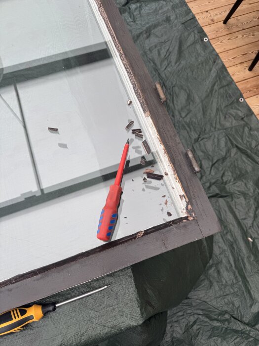 Fönsterkarm skrapas rent med röd- och blåhandtagig skruvmejsel, träflisor skrapade bort, grön presenning under.