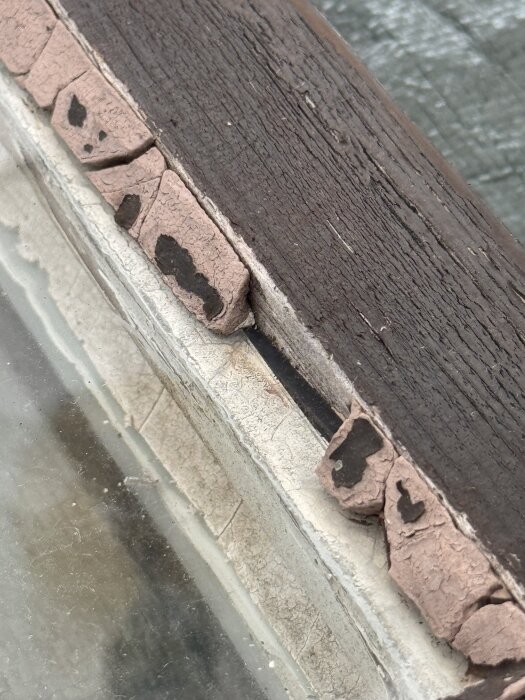 Skadat fönsterkitt på brunt träfönster med flagande färg som behöver repareras, synligt både glas och trä.