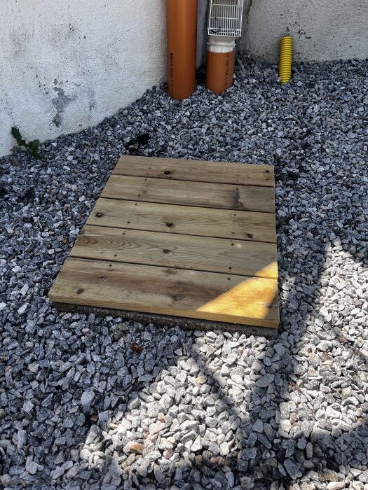 Träplattform byggd av plankor, placerad på grus vid en husvägg med orange rör och en gul slang i bakgrunden.
