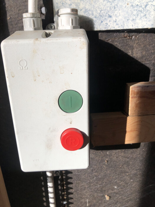 Grå kopplingsdosa med en grön och en röd knapp syns monterad på en vägg.