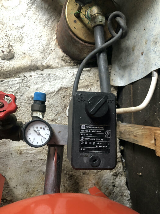 Svart elektrisk kontaktbox med märket Telemecanique och en tryckmätare i en källare, kopplad till rör och ventiler.