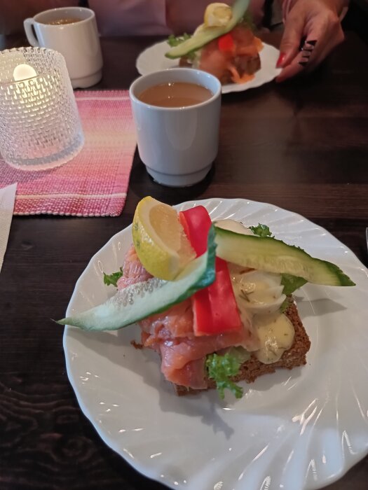 Laxsmörgås med citron och grönsaker på en vit tallrik, tillsammans med en kopp kaffe och ett ljus på bordet.
