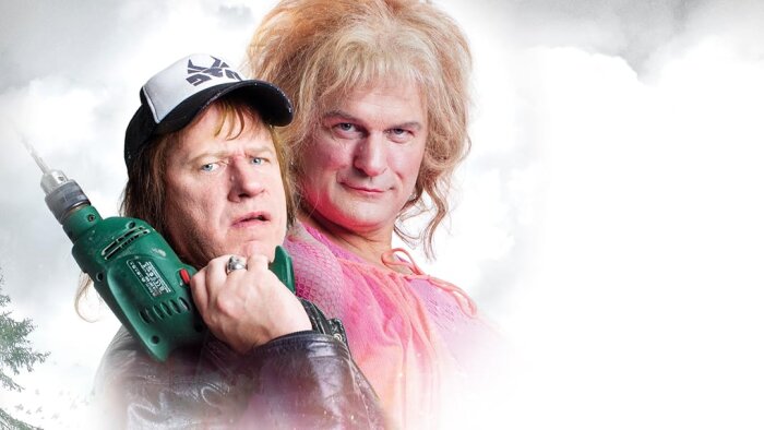 Två personer poserar humoristiskt, en med borrmaskin och keps, den andra med blont hår och rosa klänning, mot en molnig bakgrund.