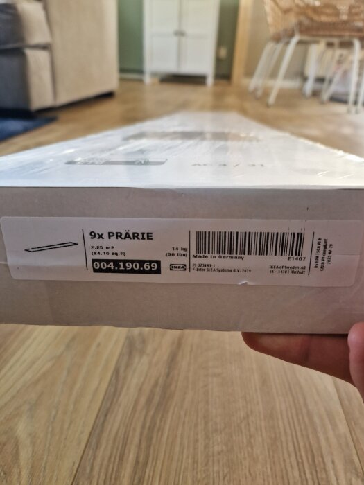 Förpackning av IKEA-golvet "Prärie" med etikett och streckkod synlig, liggande på ett trägolv i ett rum med möbler i bakgrunden.
