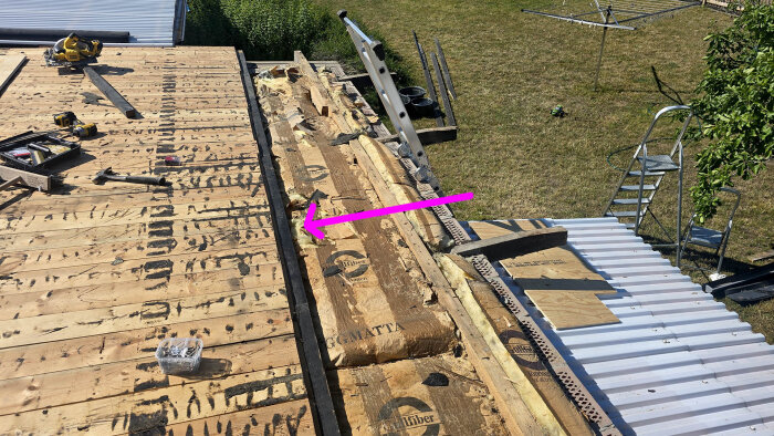 Takrenovering pågår, träpanel och verktyg syns. Isolering och byggmaterial blottat, med en stege vid sidan av taket mot gräsmattan.