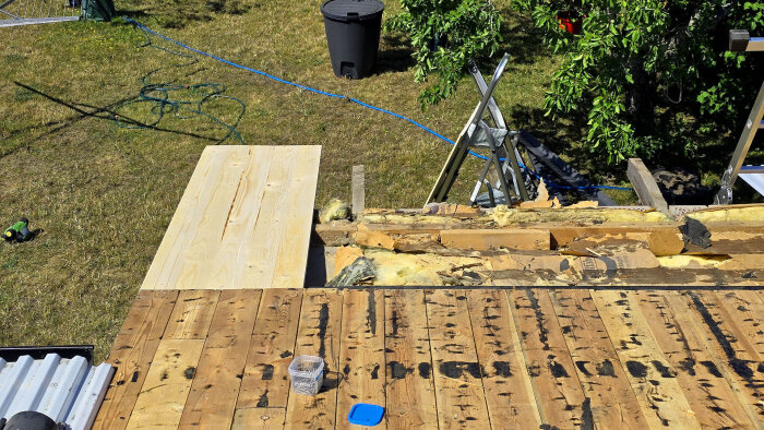 Träplankor och isolering på ett tak under renovering, med olika verktyg och material runt omkring i solen.