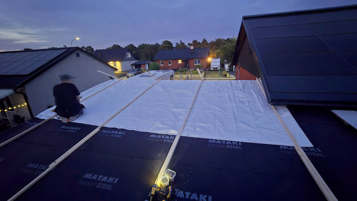 Takbyggare i arbete kvällstid, monterar takduk på en byggställning med utsikt över villakvarter.