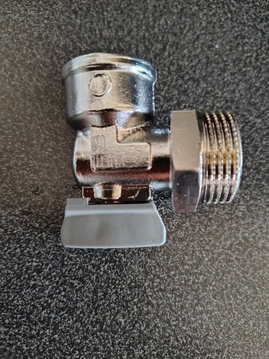 Vinkelkoppling i metall med synlig gänga, liggandes på en mörk yta, kanske för tvättmaskininstallation.