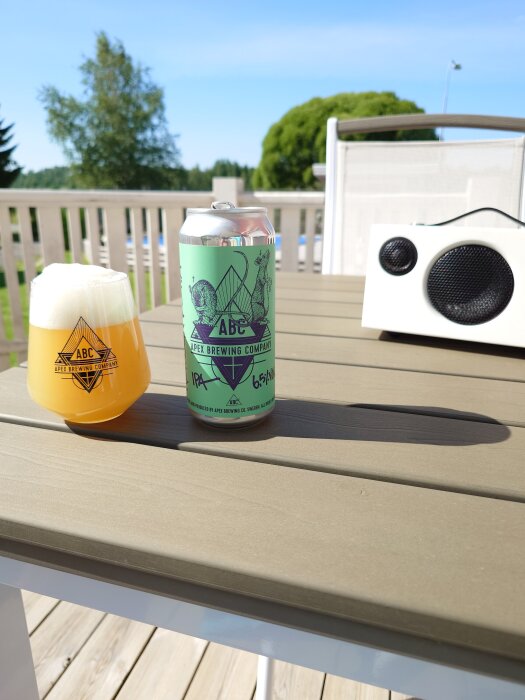 En burk och ett glas öl från Apex Brewing Company på ett bord i solen, med en bärbar högtalare i bakgrunden på en altan.