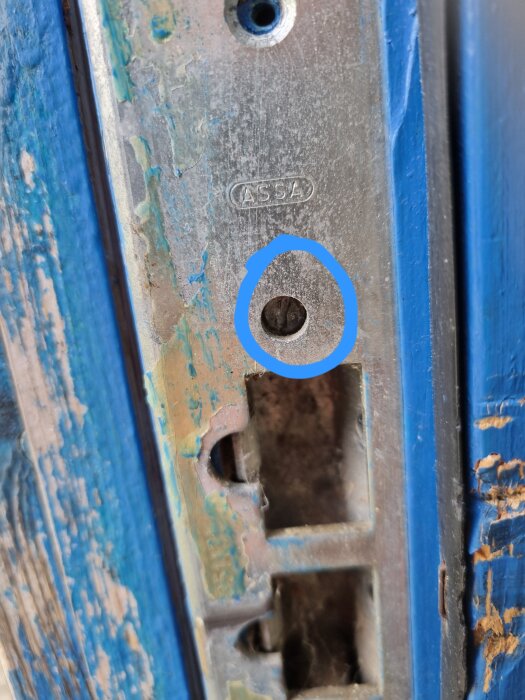 Närbild på ett dörrslutbleck av märket ASSA med synliga skruvhål, där en av de två spårskruvarna är markerad med en cirkel.