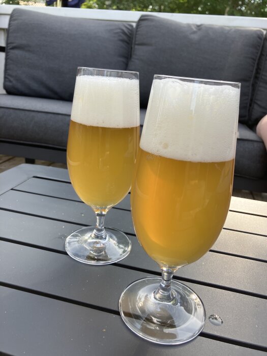 Två glas med ljus öl och skum ovanpå, placerade på ett grått bord utomhus med en soffa i bakgrunden.