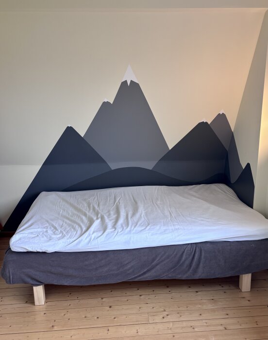 Målade bergssilhuetter i grå toner på en fondvägg bakom en säng med trägolv.