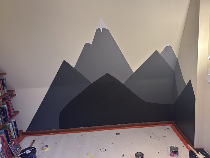 Väggmålning av berg i mörka nyanser i rum med hyllor fyllda av böcker till vänster och golvet täckt med skyddspapper och färgburkar.