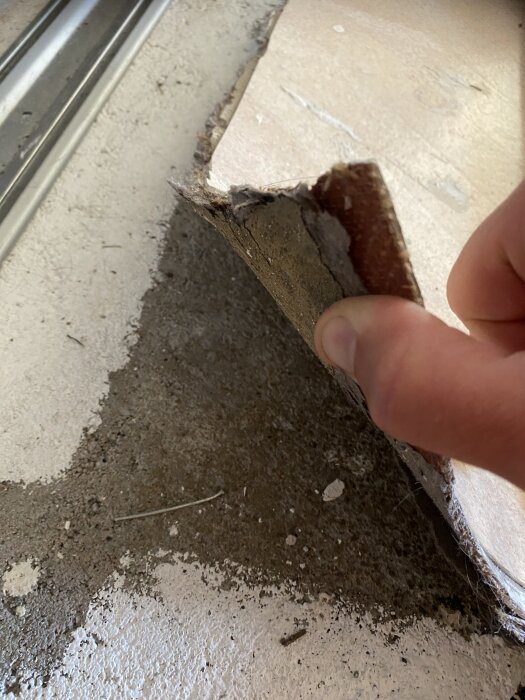 En hand drar upp en bit av gammalt golv; lagen visar linoleum på väv med svart underlag.