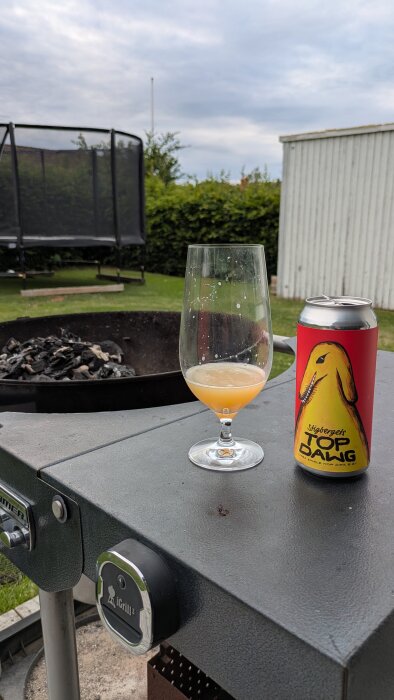 Ölglas med Top Dawg-öl på ett grillbord, bredvid en ölburk. I bakgrunden syns en nedsläckt eldplats och en studsmatta.