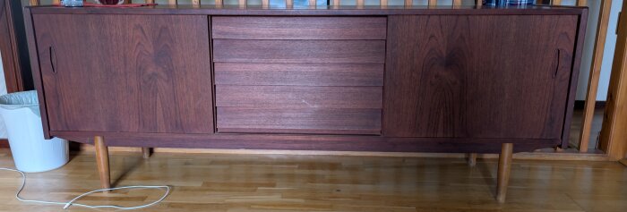 Teakmöbel med mörkt trä, skåpets mittdel har horisontella paneler, omgiven av två dörrar med integrerade handtag. Placeras på träben.