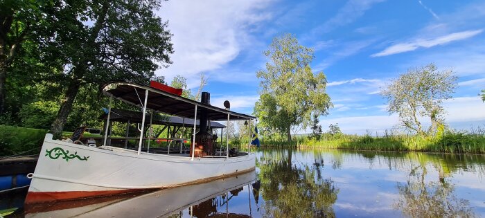 Vit båt med svensk flagga i en lugn sjö, omgiven av grönska och träd under en klarblå himmel.