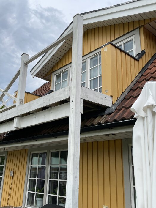 Balkong med två vita stolpar mot ett gult husfasad, en stolpe med synlig röta högst upp, framför röd takpanna och vita fönster.