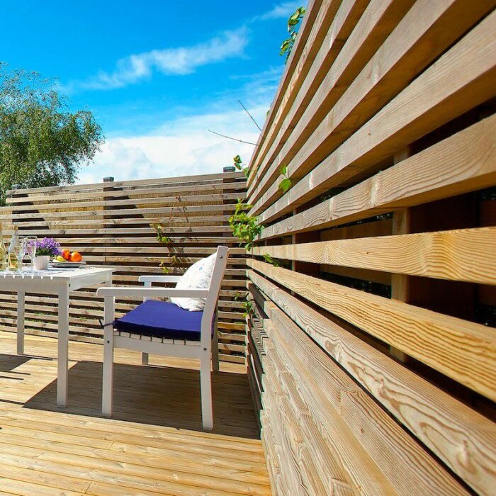 Altan med träplank som inhägnad, vit bord och stol med blå dyna, solig dag och gröna växter i bakgrunden.