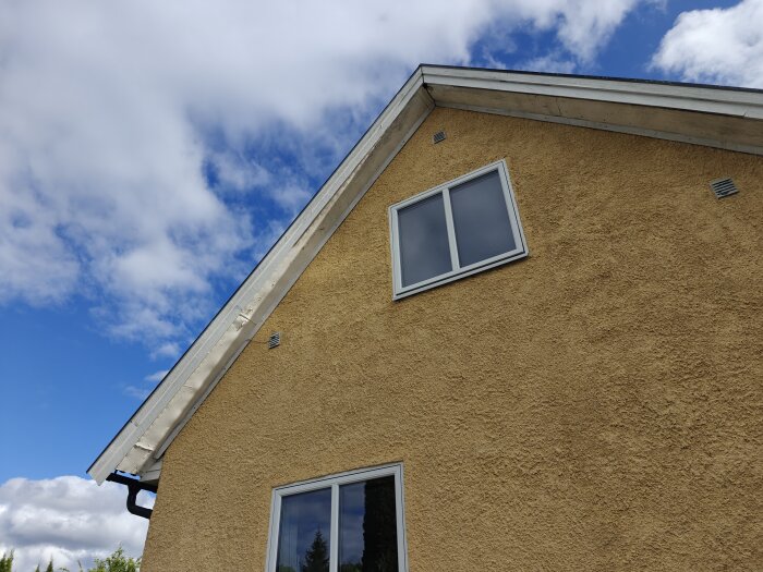 Gul husgavel med vit takfot och fönster, under klarblå himmel med vita moln.