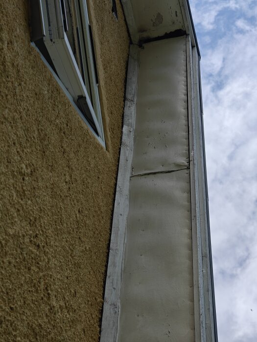 Skadad masonit och vindskivor på takfotens undersida, med fågelbo och tecken på väderslitage längs gavelsida mot himlen.