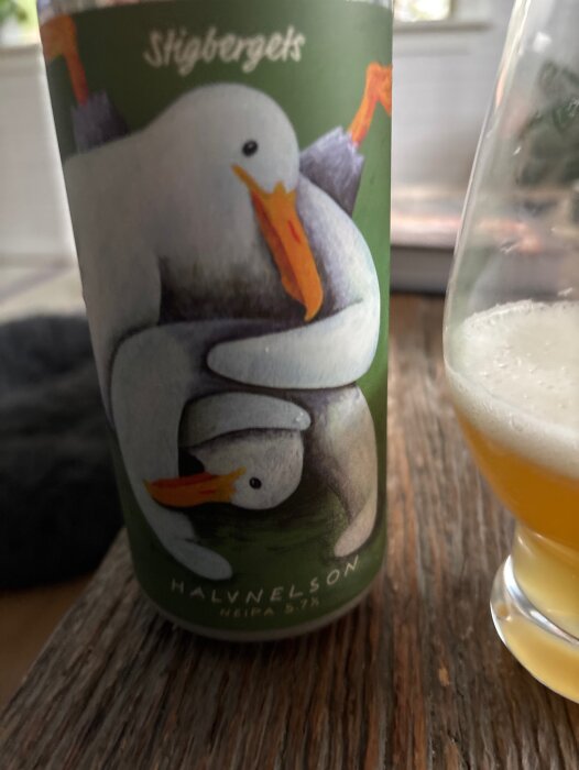 Burk med Stigbergets Halvnelson NEIPA 5,7%, motiv av två fåglar, samt ett glas med samma öl på ett träbord.