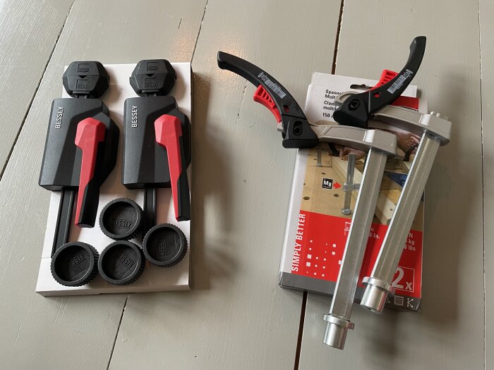 Två olika skruvtvingar på ett bord; den vänstra har svarta handtag med röda detaljer, den högra har svarta och röda detaljer och är i metall.